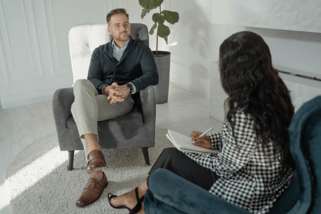 Sesja psychoterapii w Warszawie - mężczyzna w okularach siedzi w szarym fotelu naprzeciwko psychoterapeutki w jasnym gabinecie z roślinami